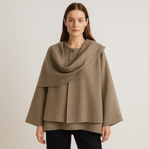 May include: A tan-colored cape-style jacket with a draped collar and button closure. The jacket has wide sleeves and a layered design. The model is wearing black pants. The background is plain.