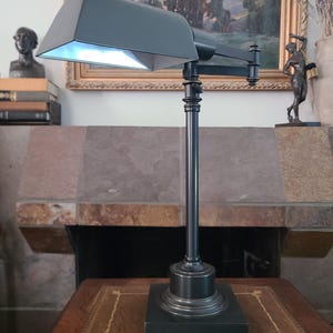 May include: A dark gray metal desk lamp featuring a rectangular base, a slender vertical post, and an adjustable horizontal arm. The lamp has a wide, trapezoidal shade that directs light downward, with a visible white interior surface.