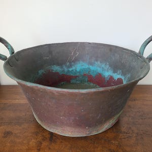 May include: A large, antique copper bowl with two arched handles. The bowl has a wide, rounded shape with a weathered patina, showing shades of brown, teal, and red. The handles are a darker teal color. The bowl sits on a wooden surface.