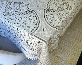 May include: A white lace tablecloth with intricate floral and geometric patterns covers a table. The tablecloth has a scalloped edge with small tassels. The table is set on a light-colored rug.