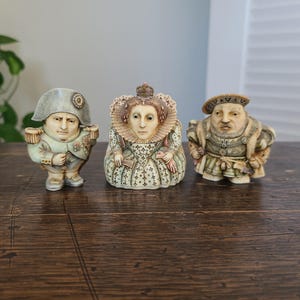 May include: Three small, detailed resin figurines depicting historical figures. The left figure wears a military uniform and bicorne hat. The center figure wears a high-collared gown and crown. The right figure wears a doublet and flat cap.