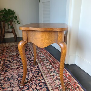 May include: A small, antique wooden side table with a curved apron and cabriole legs. The table has a light brown finish and sits on a patterned rug. The table is approximately 24 inches tall.