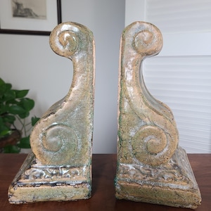 May include: A pair of ceramic bookends featuring a scroll-like design with a glossy, mottled green and tan glaze. Each piece has a wide, rectangular base and a curved, upright structure with textured detailing along the bottom edge.