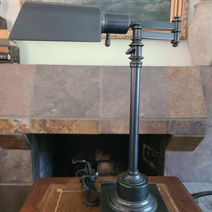 May include: A dark bronze-toned metal desk lamp featuring a rectangular shade, an adjustable swing arm, and a vertical post with decorative detailing. The lamp stands on a square base and includes a black power cord with a plug attached to the side.