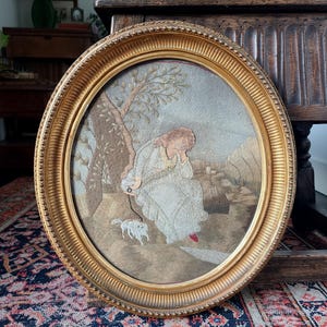 May include: An oval framed embroidery featuring a woman in a white dress with a small dog on a leash. The artwork is set in a landscape with a tree and a building, framed by a gold-colored, ornate frame.