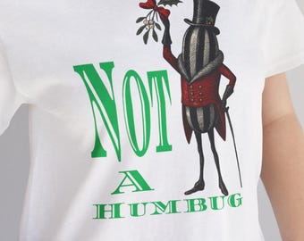 Könnte beinhalten: Weißes T-Shirt mit einem grünen Aufdruck mit der Aufschrift "NOT A HUMBUG". Die Grafik zeigt eine Zeichentrickfigur mit Zylinder und rotem Mantel, die Mistelzweige hält. Das Shirt ist aus weichem Material.