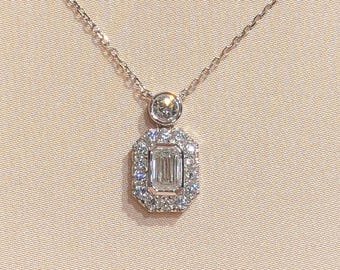 May include: A silver necklace featuring a rectangular pendant with a large, clear, emerald-cut stone, encircled by smaller, clear stones. The necklace is presented in a beige jewellery box.