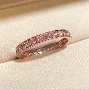 May include: Rose gold ring with a band of pink gemstones. The ring is set in a box with a cream-colored interior. The ring is a full eternity band, with pink gemstones set all the way around the band.