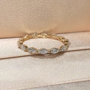 May include: A gold-colored ring with a continuous band of oval-shaped, clear gemstones. The ring is set against a soft, peach-colored background, suggesting a jewelry box or display.