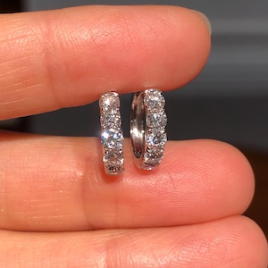 May include: A pair of small, silver hoop earrings. Each earring is encrusted with multiple small, clear gemstones. The earrings are held between two fingers, showcasing their size and design. The background is blurred, focusing attention on the jewelry.