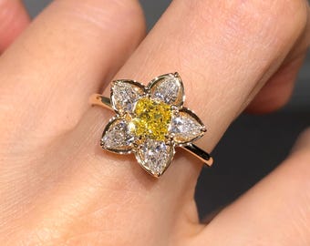 May include: A gold ring featuring a central square-cut yellow gemstone surrounded by five pear-shaped white gemstones arranged in a flower-like pattern. The band is thin and polished gold, creating a floral design that sits prominently on the finger.