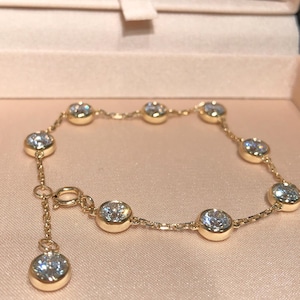 Puede incluir: Una pulsera dorada con piedras preciosas redondas y claras engastadas en biseles individuales. La pulsera se exhibe en una caja de joyería rosa, mostrando su delicada cadena y cierre.