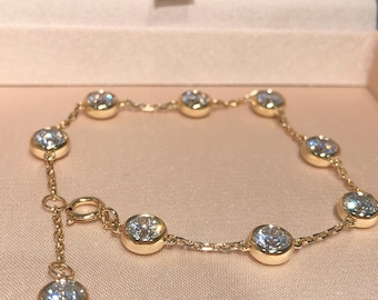 Puede incluir: Una pulsera dorada con piedras preciosas redondas y claras engastadas en biseles individuales. La pulsera se exhibe en una caja de joyería rosa, mostrando su delicada cadena y cierre.