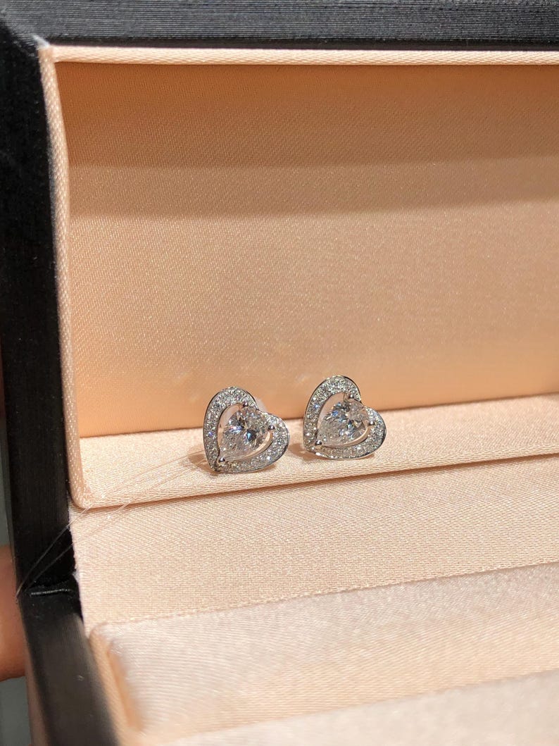 May include: A pair of heart-shaped stud earrings. Each earring features a central, clear gemstone surrounded by smaller, clear stones. The earrings are set in a silver-colored metal and rest on a peach-colored fabric inside a black box.