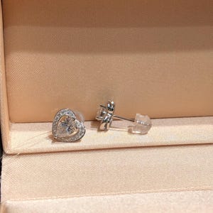 May include: A pair of silver stud earrings. One earring is a heart shape with a clear gemstone center and a halo of smaller stones. The other earring is visible with a post and backing. The earrings are displayed in a pink jewelry box.