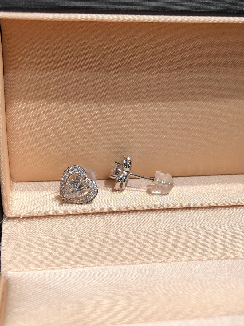 May include: A pair of silver stud earrings. One earring is a heart shape with a clear gemstone center and a halo of smaller stones. The other earring is visible with a post and backing. The earrings are displayed in a pink jewelry box.