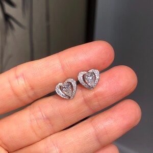 May include: A pair of heart-shaped stud earrings. Each earring features a central, clear gemstone surrounded by smaller, clear stones, all set in a silver-colored metal. The earrings are designed with a delicate, romantic aesthetic.