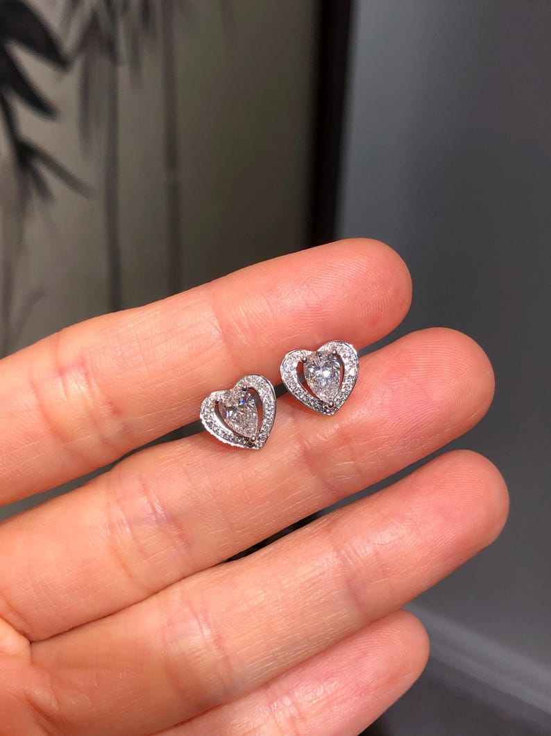 May include: A pair of heart-shaped stud earrings. Each earring features a central, clear gemstone surrounded by smaller, clear stones, all set in a silver-colored metal. The earrings are designed with a delicate, romantic aesthetic.