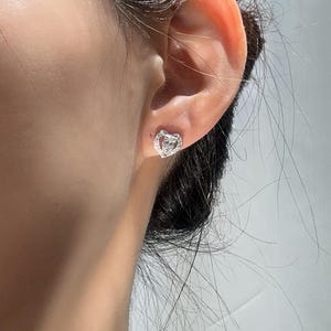 May include: A close-up of a silver heart-shaped stud earring with a small diamond in the center. The earring is worn on an ear. The earring is surrounded by smaller diamonds.