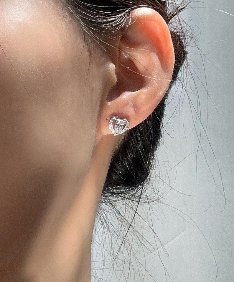 May include: A close-up of a silver heart-shaped stud earring with a small diamond in the center. The earring is worn on an ear. The earring is surrounded by smaller diamonds.