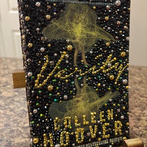 May include: A hardcover book with a black cover encrusted with black, gold, and silver rhinestones. The cover features a yellow illustration of a person and the author's name, Colleen Hoover, in gold lettering. The book is displayed on a wooden stand.