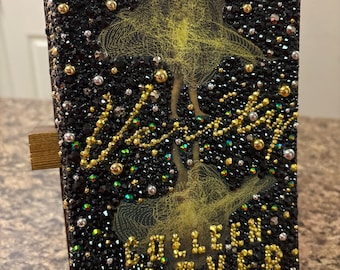 May include: A hardcover book with a black cover encrusted with black, gold, and silver rhinestones. The cover features a yellow illustration of a person and the author's name, Colleen Hoover, in gold lettering. The book is displayed on a wooden stand.