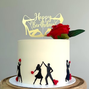 May include: A cylindrical cake with smooth white frosting, decorated with black glitter silhouettes of dancing couples and small red flowers. A gold cake topper reads Happy Birthday above two high-heeled shoes, with a large red rose attached.