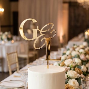 May include: A three-tiered white wedding cake with a gold monogram cake topper. The cake sits on a table set for a wedding reception, with floral arrangements and crystal glassware.