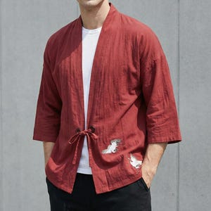 Op de afbeelding: Rode jas in kimono-stijl met driekwart mouwen en een striksluiting. De jas heeft geborduurde kraanvogelontwerpen bij de zoom. De persoon draagt er een wit T-shirt onder en een zwarte broek.