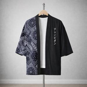May include: A black and white kimono-style jacket with a paisley pattern on the left side and solid black on the right. The right side features white Japanese text. The jacket is hanging on a clothing rack.