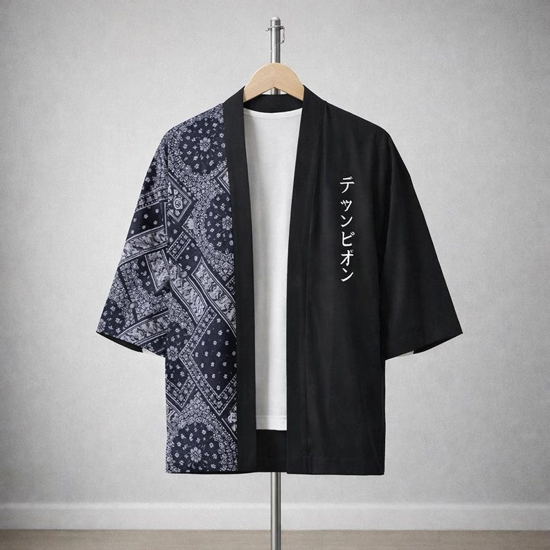 May include: A black and white kimono-style jacket with a paisley pattern on the left side and solid black on the right. The right side features white Japanese text. The jacket is hanging on a clothing rack.