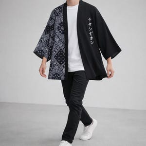 May include: Black and navy blue kimono-style jacket with a paisley pattern on the left side and Japanese text on the right. The jacket is worn over a white t-shirt and black pants. The person is wearing white sneakers.