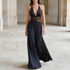 May include: A black halter-neck maxi dress with a plunging neckline and a lace panel at the waist. The dress has a flowing skirt and is made of a silky material. The model is walking in a well-lit corridor.