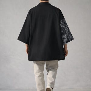 May include: Black kimono-style shirt with a contrasting paisley pattern on one sleeve. The shirt has a relaxed fit and three-quarter sleeves. The model is wearing light-colored pants and white shoes.