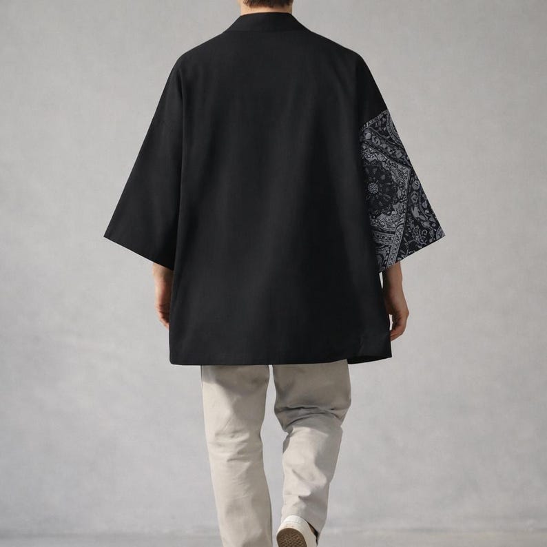 May include: Black kimono-style shirt with a contrasting paisley pattern on one sleeve. The shirt has a relaxed fit and three-quarter sleeves. The model is wearing light-colored pants and white shoes.