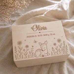 Op de afbeelding: Een rechthoekige, lichtgekleurde houten doos met een lasergravure van een slapend reekalfje, wilde bloemen, een vlinder en sterren. De tekst luidt Olivia, 2026.06.21, 10:10, 3650 g en 55 cm, met een klein hartje onder de naam.