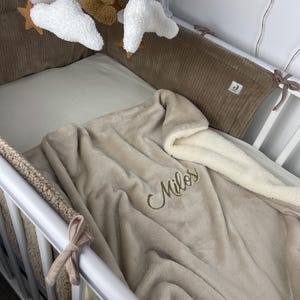 Könnte beinhalten: Ein Babybett mit einer weichen, beigen Decke, auf der der Name "Milos" in goldfarbenem Faden gestickt ist. Das Bett hat eine braune Cord-Bettumrandung und ein Mobile mit Wolken, Sternen und einem Teddybären.