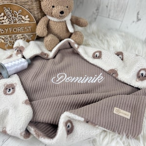Op de afbeelding: Een bruine gebreide babydeken met een witte sherpa voering en een herhalend patroon van berengezichtjes. De deken is geborduurd met de naam Dominik in wit schrift en bevat een klein rechthoekig merklabel met de tekst Baby Forest.
