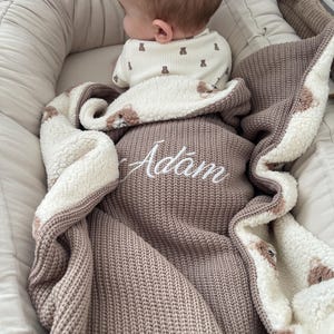 Può includere: Una morbida coperta lavorata a maglia color tortora con fodera in pile sherpa bianco e un motivo ripetuto di piccoli orsi marroni. La coperta è personalizzata con il nome Adam ricamato in corsivo bianco al centro.