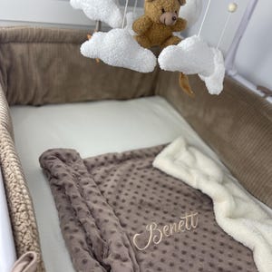 Può includere: Una culla per neonati con una giostrina con orsetto marrone e una coperta marrone ricamata con il nome "Benett". La coperta ha una trama a pois ed è appoggiata su un lenzuolo bianco. La culla è foderata in velluto a coste marrone.