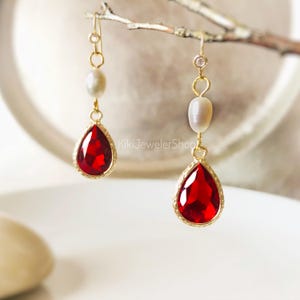 May include: A pair of gold-tone drop earrings featuring a faceted, pear-shaped red stone suspended below a smooth, oval-shaped white pearl. Each earring includes a small, clear circular accent at the top, connected by thin gold-tone metal links.