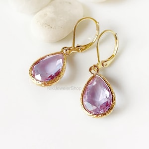 May include: A pair of gold-colored leverback earrings with teardrop-shaped, light purple gemstones. The gemstones are faceted and set in a gold-colored bezel. The earrings are displayed on a white surface.