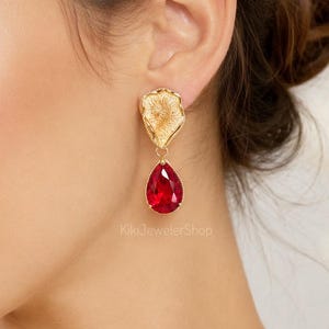 May include: A gold-toned earring featuring a textured, organic-shaped top piece connected to a faceted, pear-shaped red crystal drop. The earring is displayed on an earlobe, and the text KikiJewelerShop is visible in the lower center area.