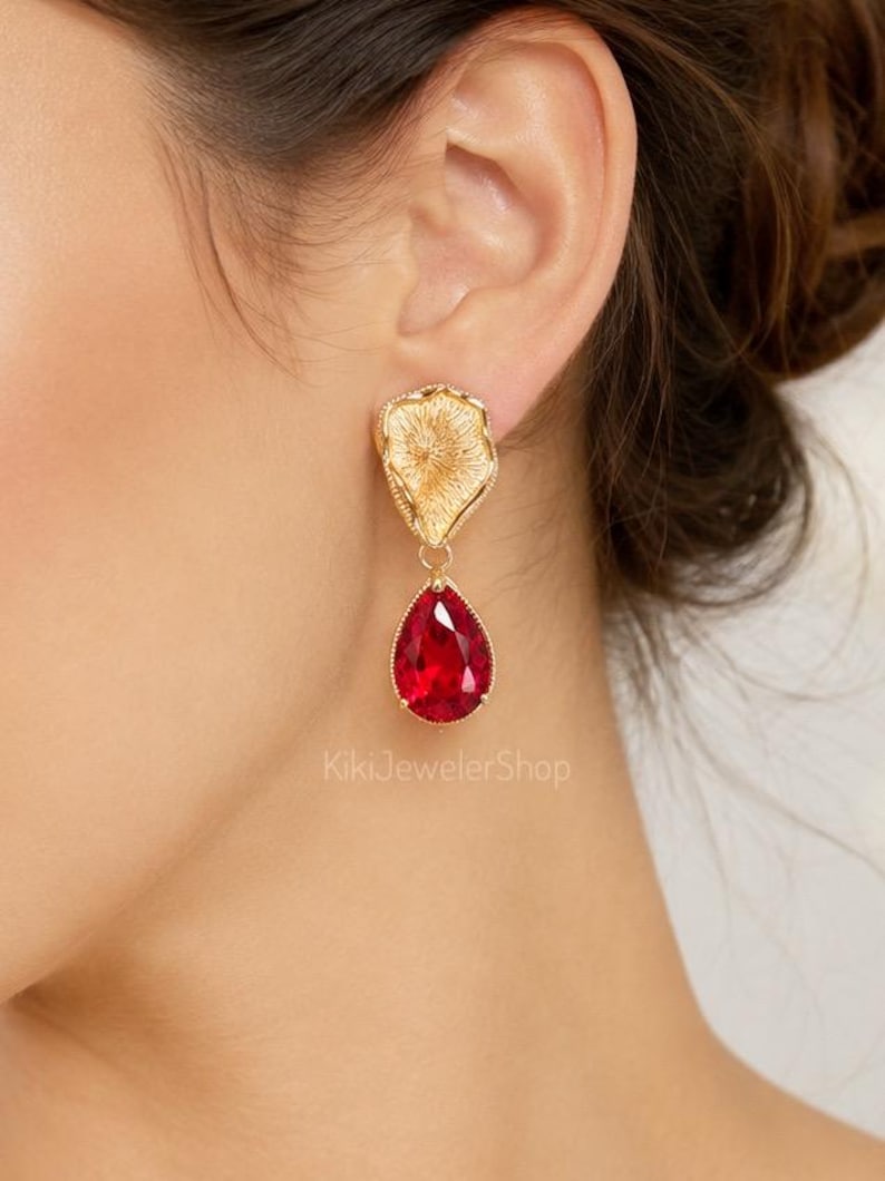 May include: A gold-toned earring featuring a textured, organic-shaped top piece connected to a faceted, pear-shaped red crystal drop. The earring is displayed on an earlobe, and the text KikiJewelerShop is visible in the lower center area.