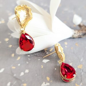 May include: A pair of gold-tone stud earrings featuring a textured, fan-shaped top piece and a dangling, faceted, pear-shaped red crystal drop. The text KikiJewelerShop is visible in the center. The earrings have a post-back closure mechanism.