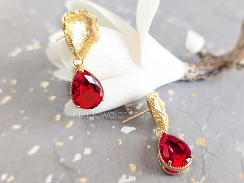 May include: A pair of gold-tone stud earrings featuring a textured, fan-shaped top piece and a dangling, faceted, pear-shaped red crystal drop. The text KikiJewelerShop is visible in the center. The earrings have a post-back closure mechanism.