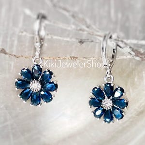 May include: A pair of silver-toned flower earrings. Each earring features a flower design with dark blue gemstone petals surrounding a central white gemstone. The earrings are suspended from a hinged lever back closure. The text "KikiJewelerShop" is visible.