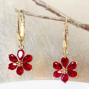 May include: A pair of gold-colored earrings, each featuring a flower-shaped pendant. The flower petals are a vibrant red, and the earrings are suspended from a hinged hoop. The earrings are set against a neutral background.