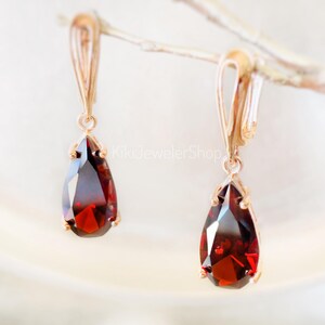 May include: A pair of drop earrings featuring faceted, pear-shaped deep red garnet gemstones set in rose gold metal. The earrings have a secure lever-back closure and the text KikiJewelerShop is visible in the center of the image.
