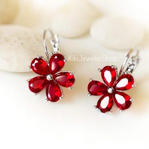 May include: A pair of silver-toned leverback earrings featuring five faceted, pear-shaped red stones arranged in a flower petal pattern. The text KikiJewelerShop is visible in the center. The earrings have a polished metallic finish and clear, vibrant red gemstone petals.
